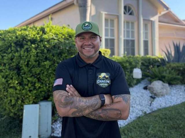 Juan Chacon, smiling with arms crossed, in his Chacon Family Lawn Care cap and polo in front of a South Florida home. – Chacon Family Lawn Care