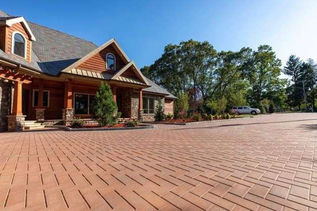 A large brick driveway leads to a wooden house with trees and a parked truck in the background. – Palm Beach Outdoor Contracting